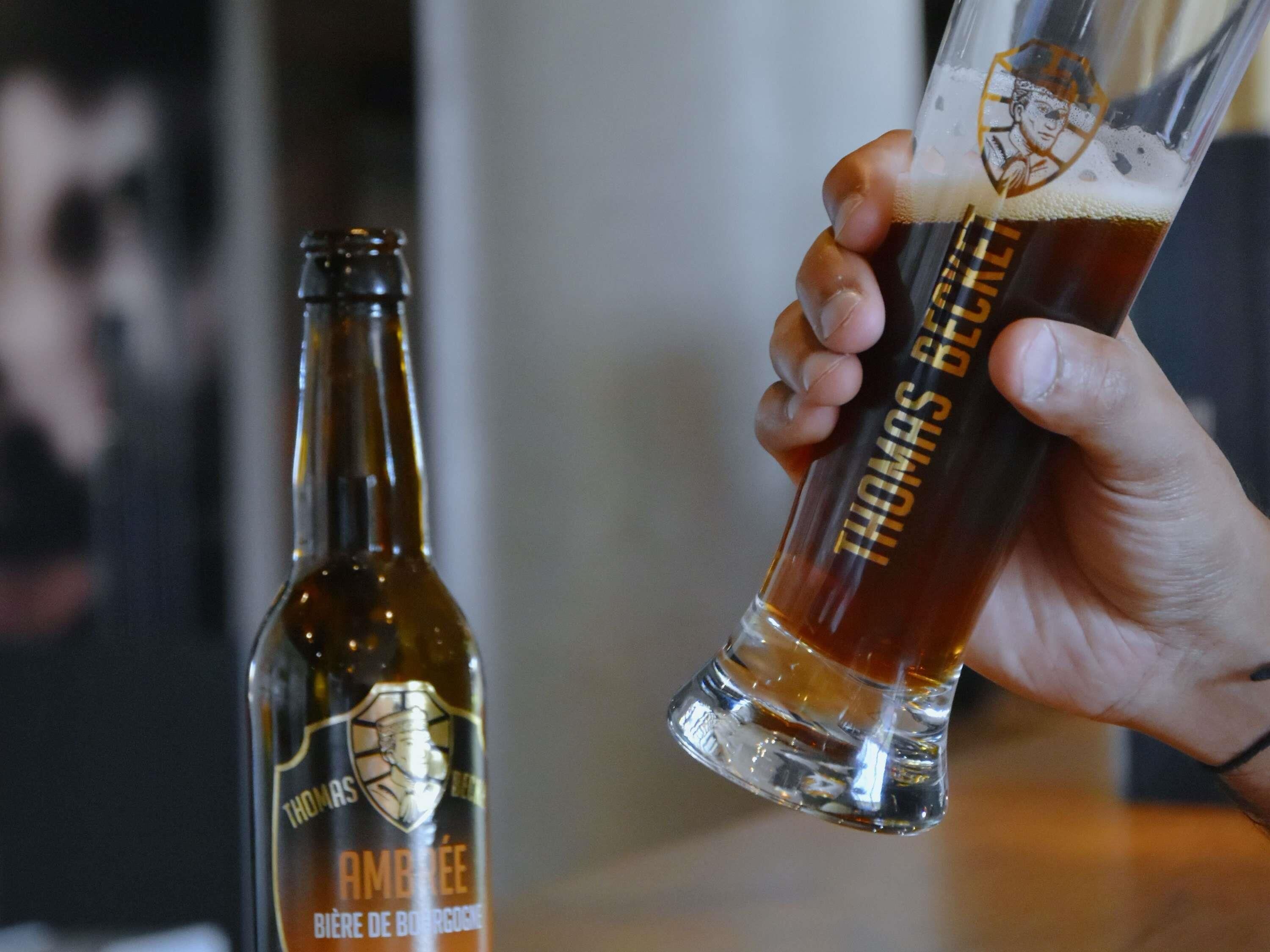 A hand holding a glass of beer with foam, and a bottle labeled 'THOMAS BRUGGOSKE' in the background.