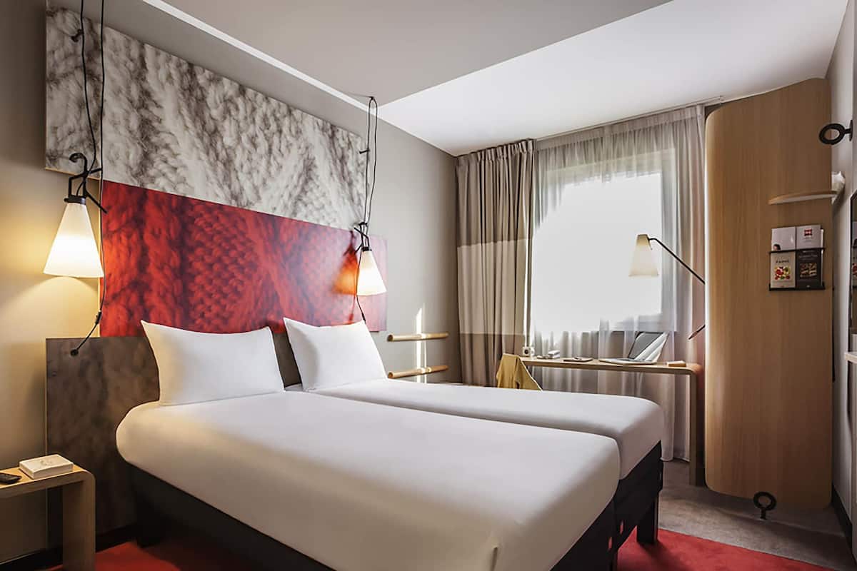 A hotel room with a bed, bedside tables, a desk, a chair, a window with curtains, and a wall with a marble-like design.