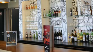 Een barruimte met flessen en glazen op planken, een poster die een drankspecial promoot, en een zwart-wit muurschildering.