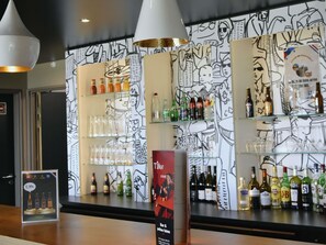 Zona de bar con estantes llenos de botellas y vasos, un cartel publicando una oferta de bebida, y un mural en blanco y negro en la pared.