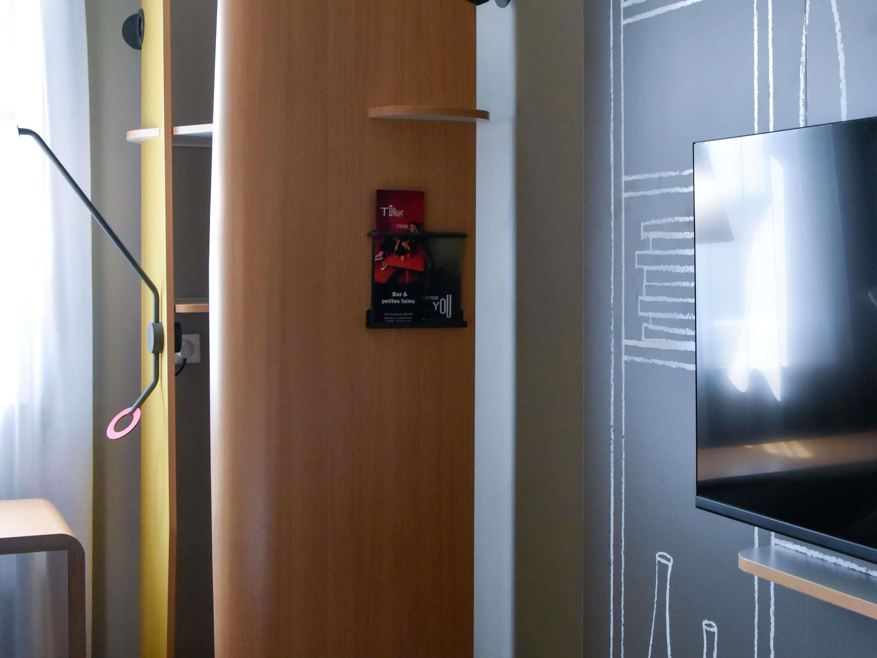 A modern hotel room with a large flat-screen TV mounted on the wall, a wooden door, and a small poster advertising a bar and restaurant.