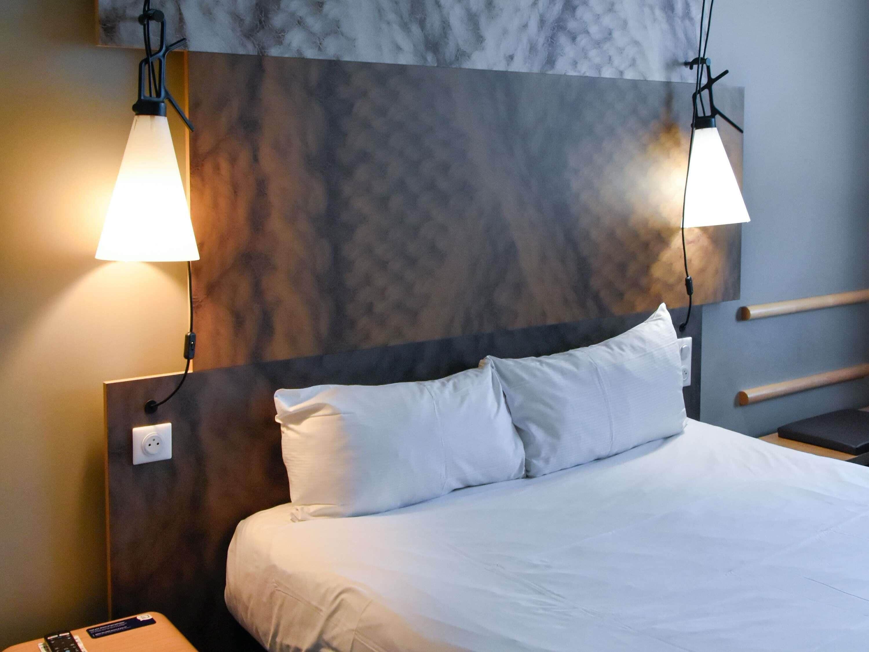 A hotel room with a marble headboard, two bedside lamps, a bed with white linens, and a bedside table with a remote control.