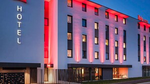 Een modern hotelgebouw met het 'ibis' logo en 'HOTEL' aanduiding, met een platte gevel met gelijkmatig verdeelde ramen en rode accentverlichting.