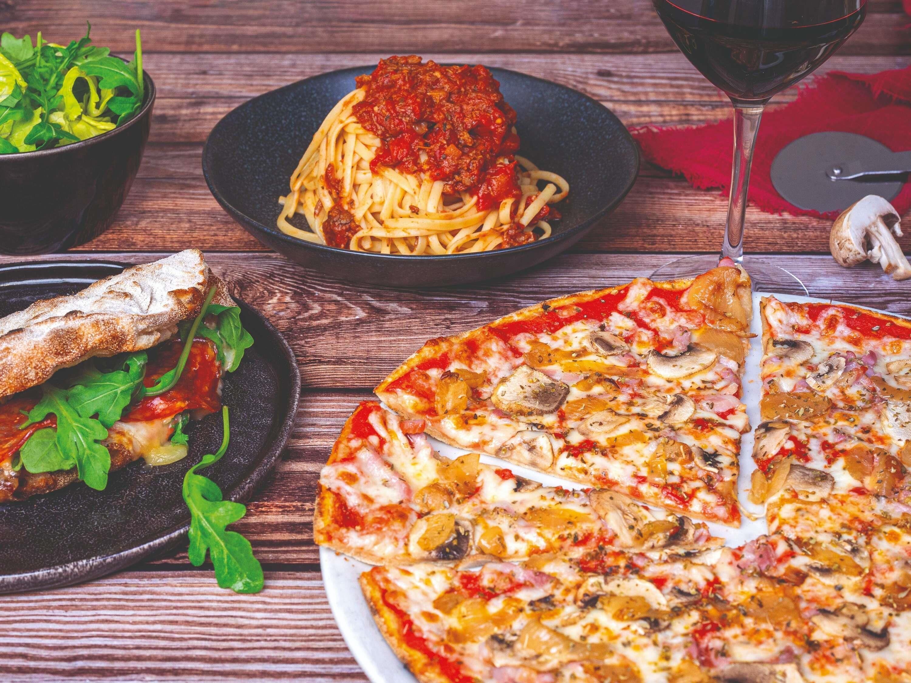 Una pizza con diversi condimenti, una ciotola di verdure, un piatto di pasta al pomodoro e un bicchiere di vino rosso su un tavolo di legno.