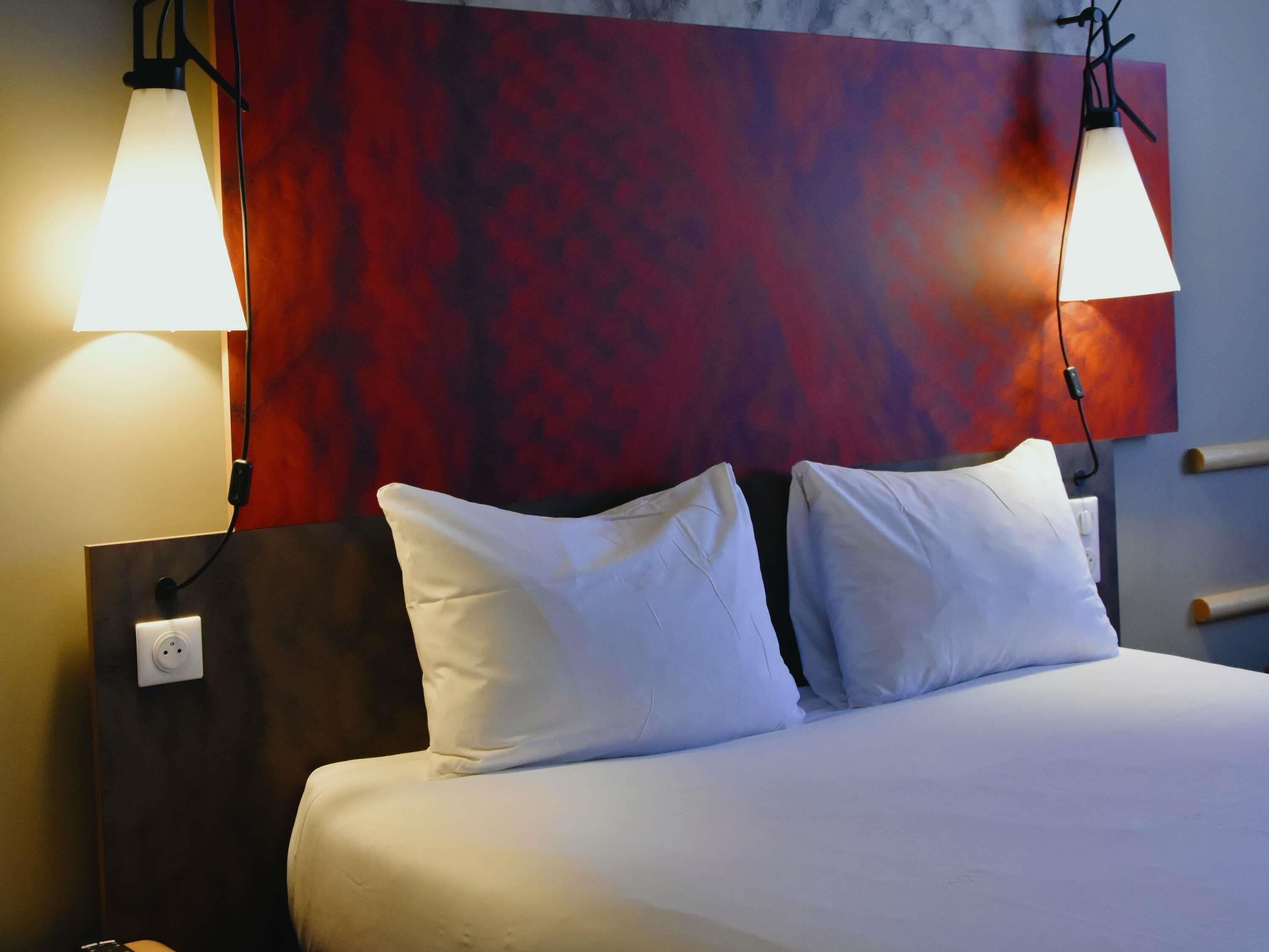 A hotel room with a bed, two white pillows, a red abstract wall, and two wall-mounted lamps.