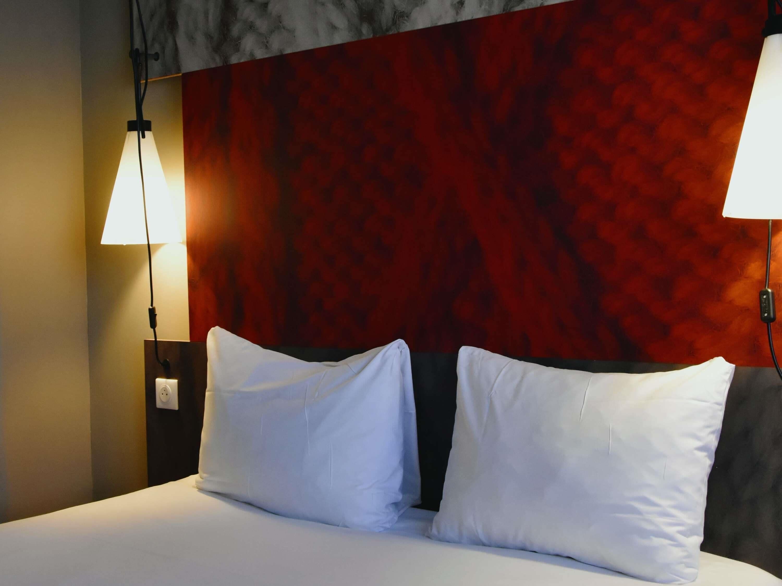Habitación de hotel con cama, dos almohadas blancas, pared texturizada roja y dos lámparas fijadas a la pared.