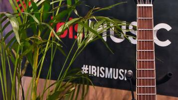 Eine Gitarre mit schwarzem Kopf und Korpus aus Holz, vor einem Hintergrund mit dem Text '#IBISMUSIC' und einem teilweise sichtbaren roten und schwarzen Design.