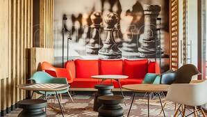 Een moderne zithoek met een rode bank, kleurrijke stoelen en een muurschildering met een schaakthema.