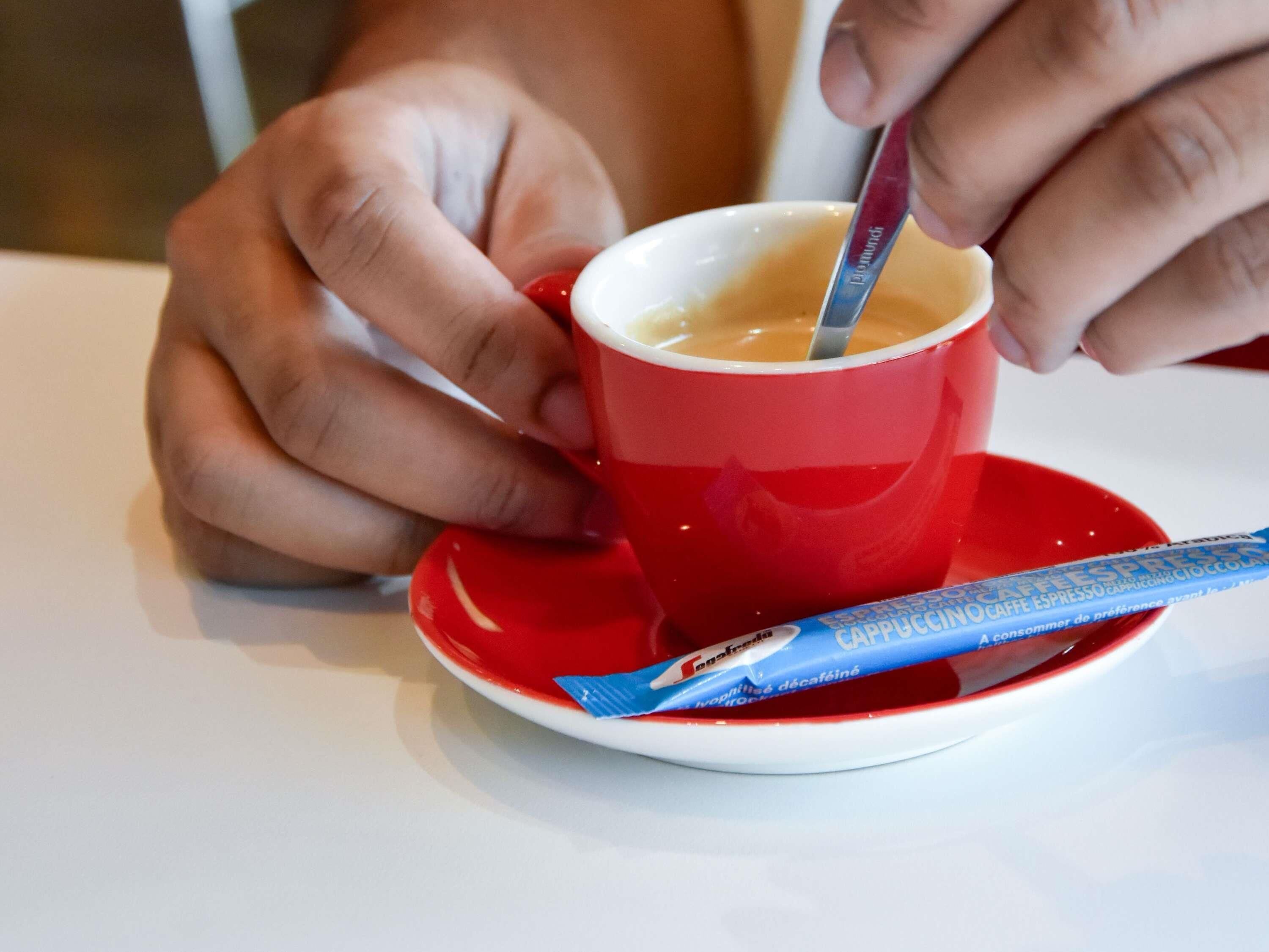 Una persona sosteniendo una taza roja con una cuchara, un sobre azul a su lado y un platito blanco sobre una mesa.