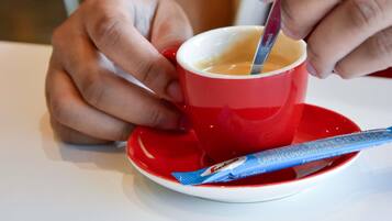 Una persona sosteniendo una taza roja con una cuchara, un sobre azul a su lado y un platito blanco sobre una mesa.