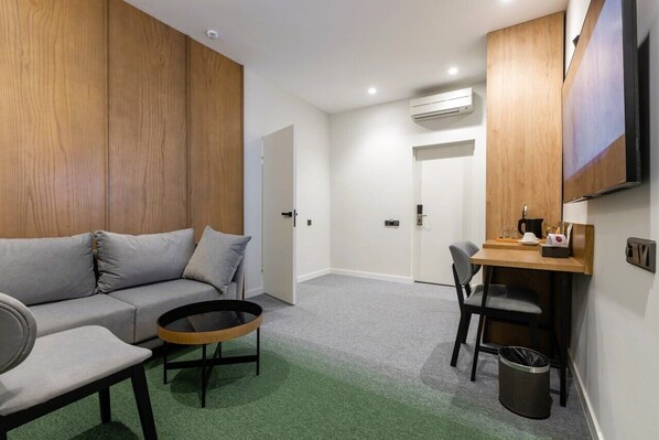 Une chambre d’hôtel moderne avec un canapé gris, une petite table ronde, un bureau en bois avec une chaise et un téléviseur fixé au mur.