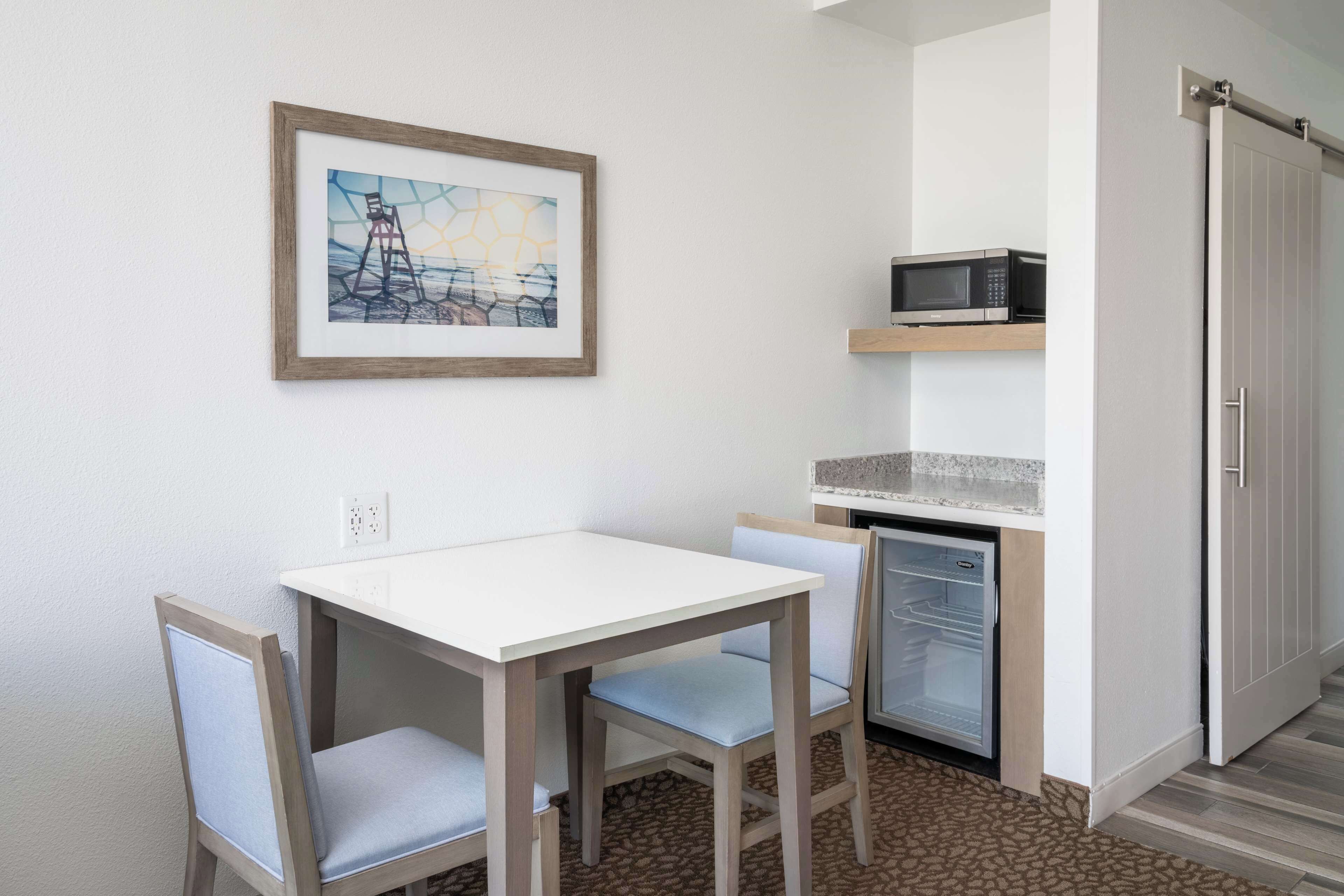 A compact room with a white table and two chairs, a small refrigerator, a microwave on a shelf, and a door with a sliding mechanism.