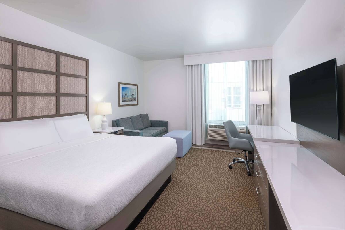 Habitación de hotel con una cama grande, un escritorio con silla, televisión y ventana con cortinas.