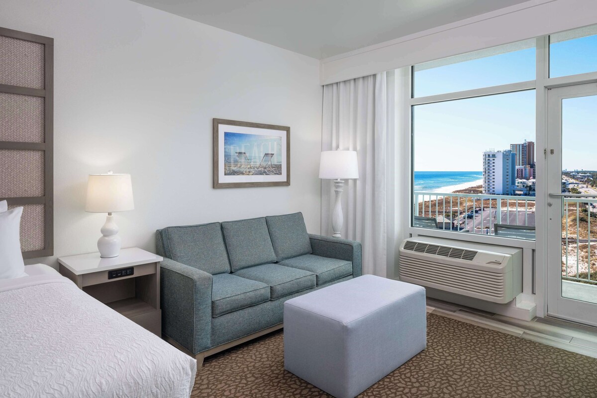 A hotel room with a bed, a grey sofa, a small ottoman, a nightstand with a lamp, a framed picture on the wall, and a large window with a view of the beach and city buildings.