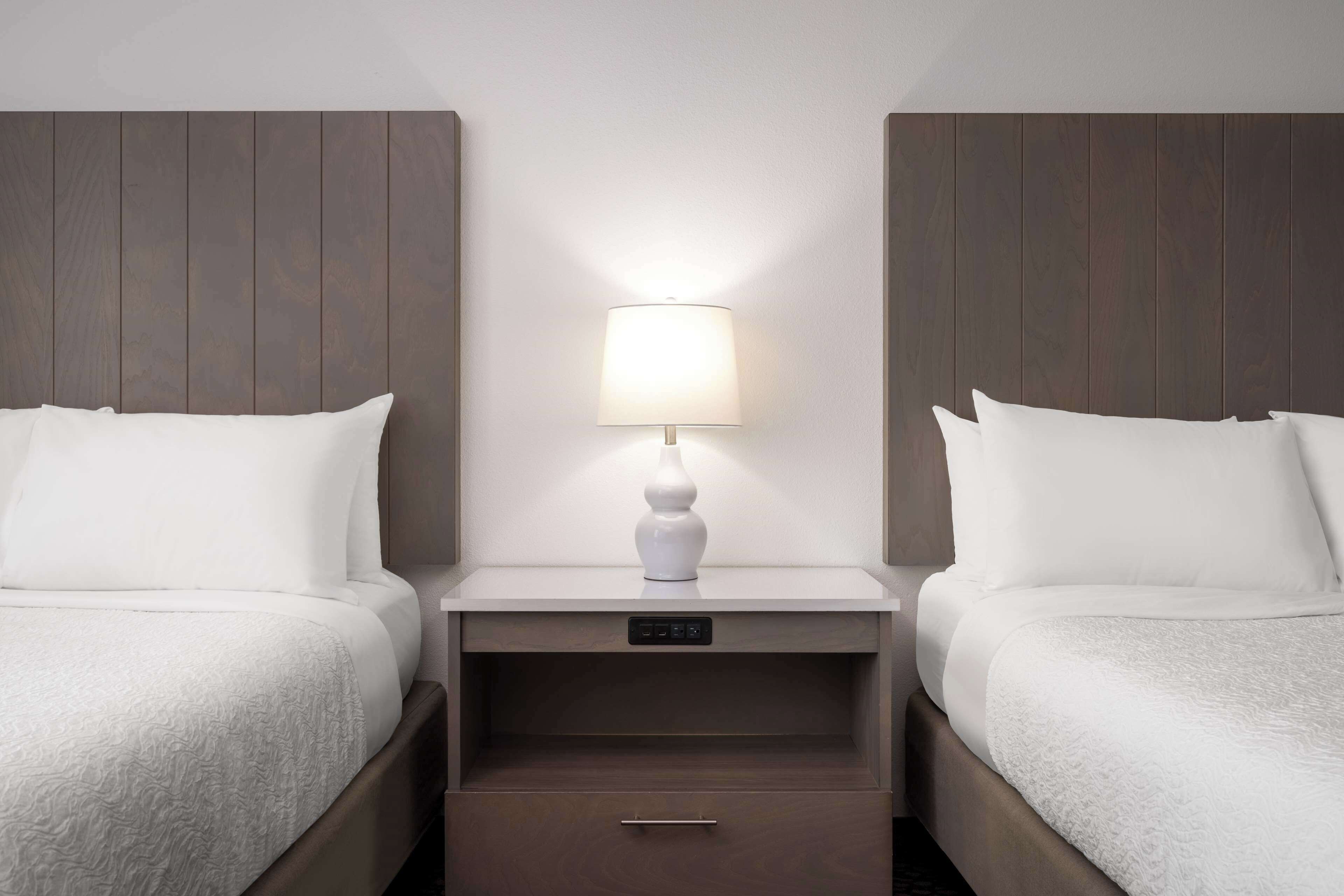 A hotel room with two beds, a bedside table with a lamp, and a control panel.