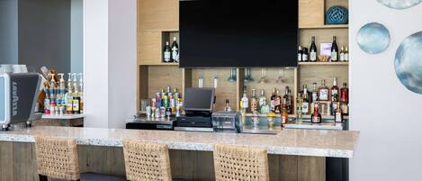 A bar area with a flat-screen TV, a cash register, and a variety of liquor bottles. There are wicker chairs and a marble countertop.