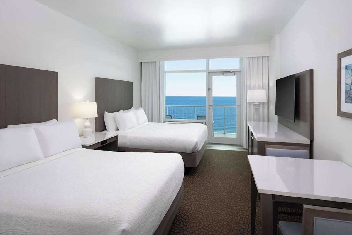 Habitación de hotel con dos camas, televisión, escritorio y balcón con vista al mar.
