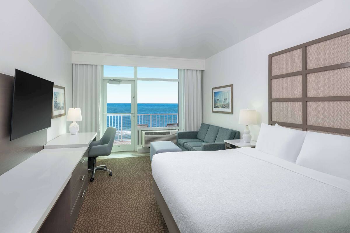 Una habitación de hotel con una cama grande, un escritorio con una silla, un sofá, un televisor y vista al océano.