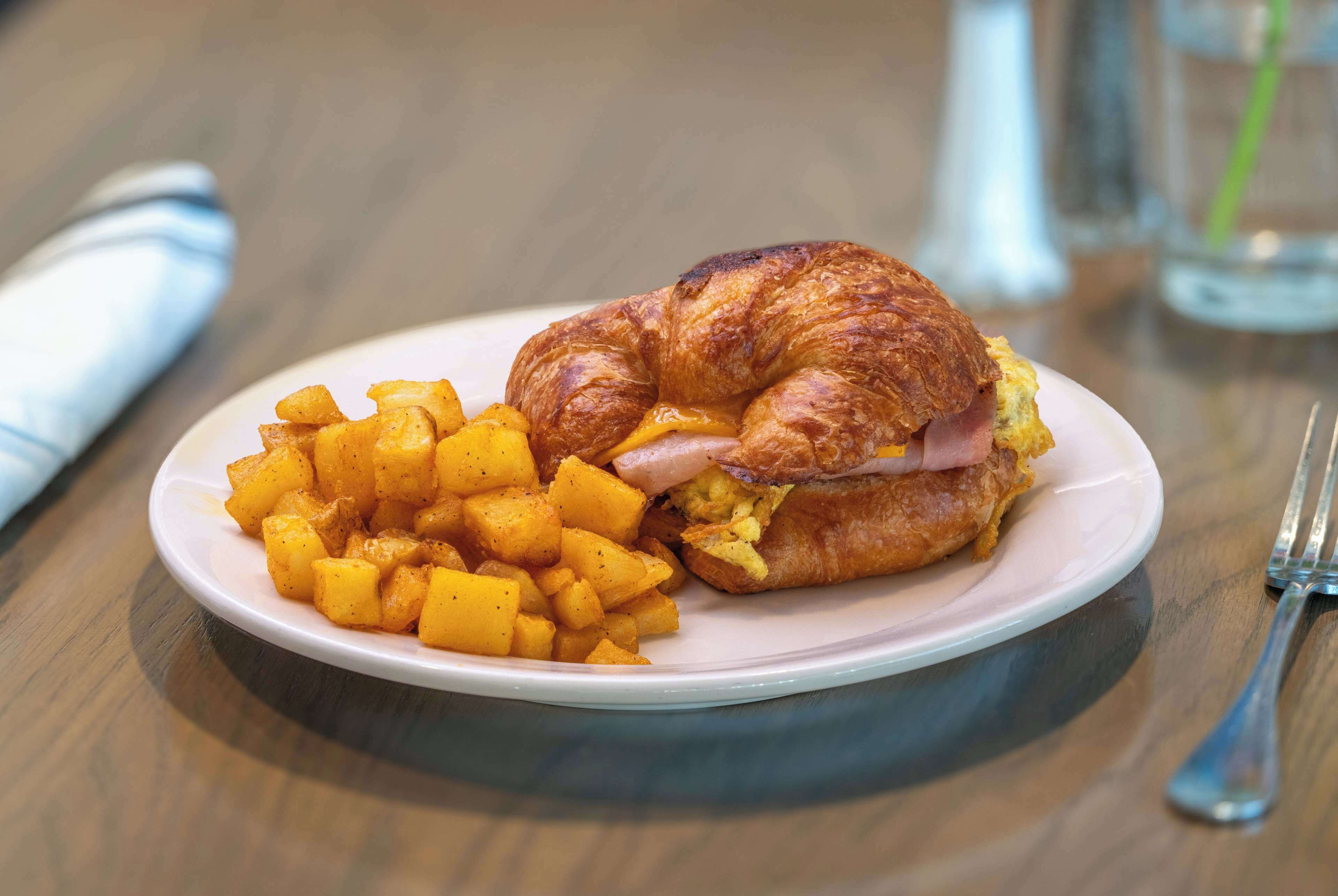 A plate with a croissant sandwich and cubed potatoes.
