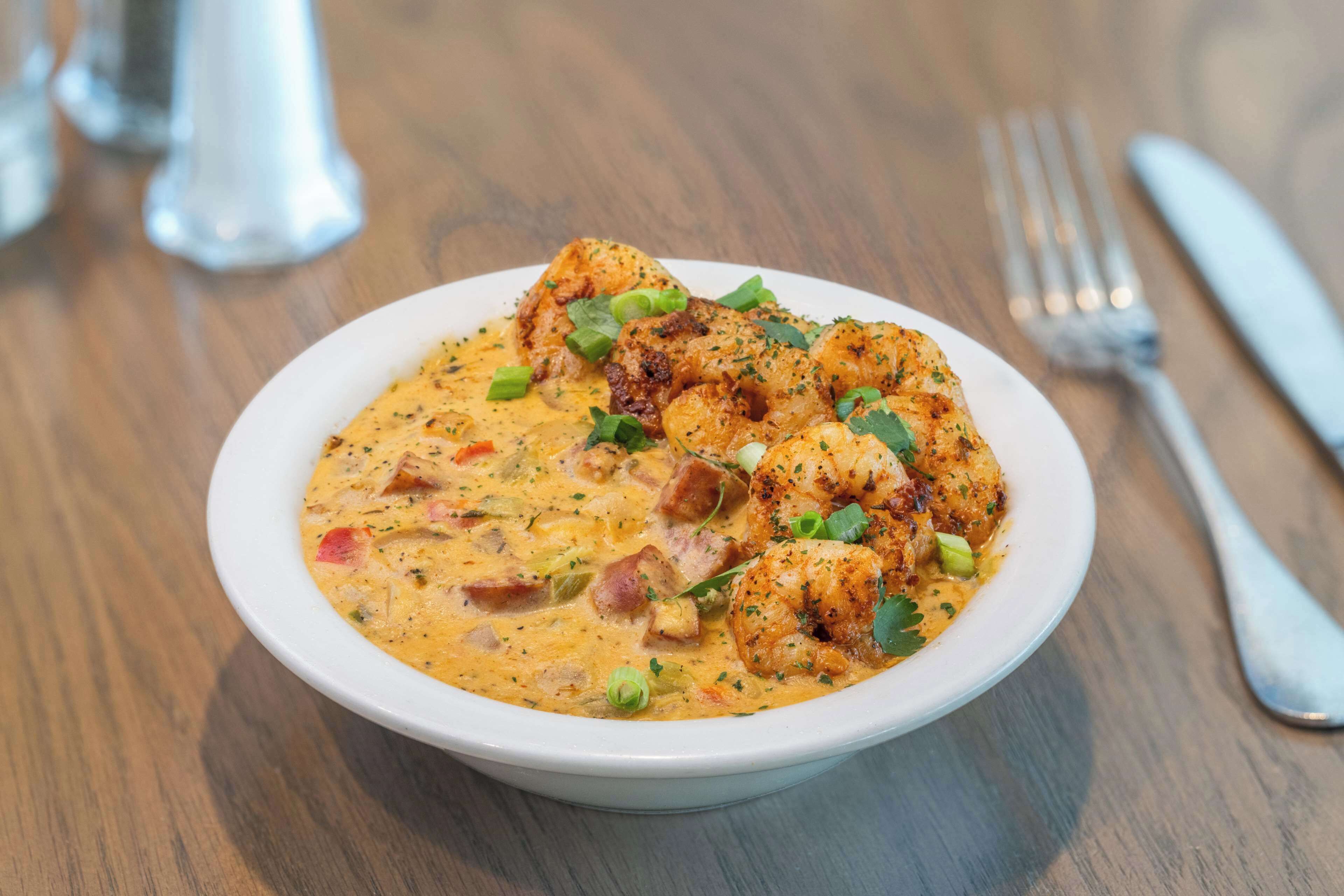 A white bowl of seafood soup with shrimp, garnished with green onions and parsley, on a wooden table with a fork and knife beside it.
