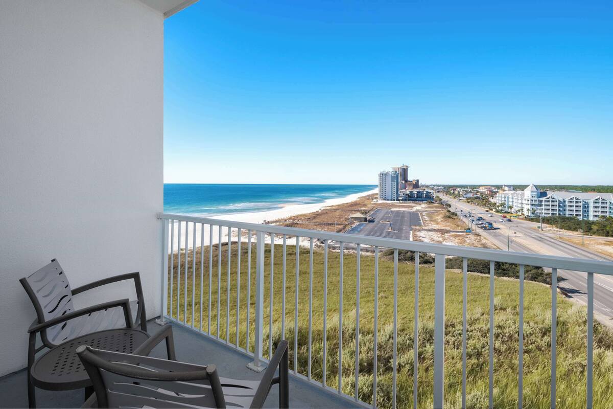 Un balcón con vista a la playa y al océano, barandilla blanca y dos sillas.