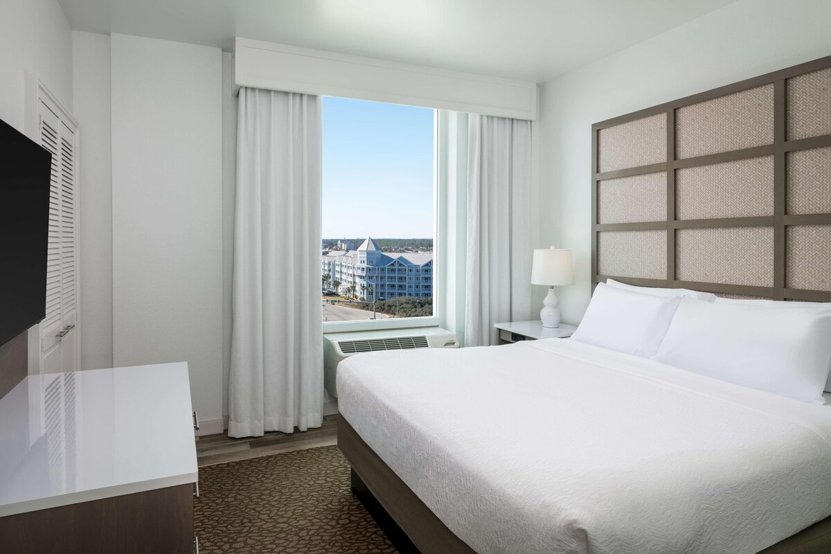 Una habitación de hotel con cama, escritorio, ventana con cortinas y vista a edificios.