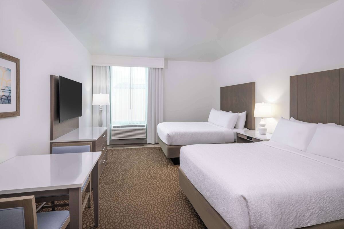 Habitación de hotel con dos camas, un escritorio, un televisor y un ventanal con cortinas.