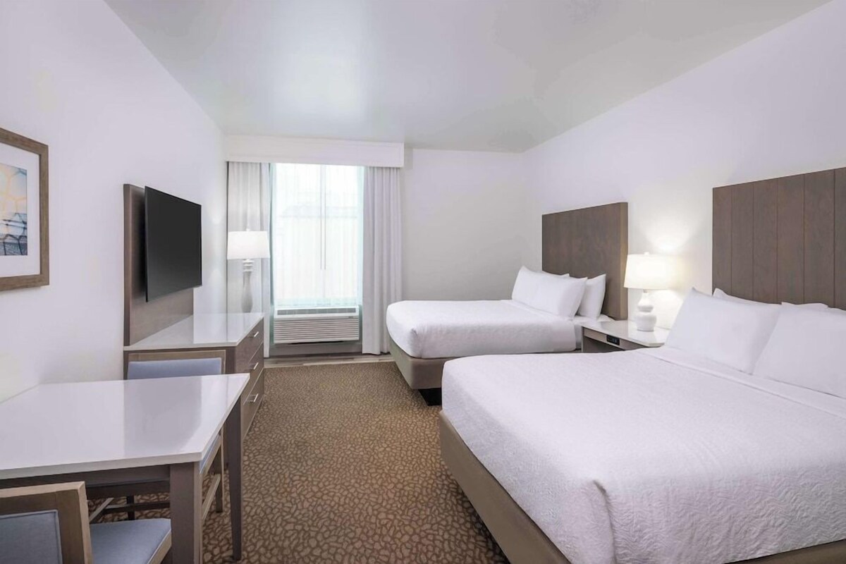 Habitación de hotel con cama, escritorio, silla, televisión y ventana con cortinas.