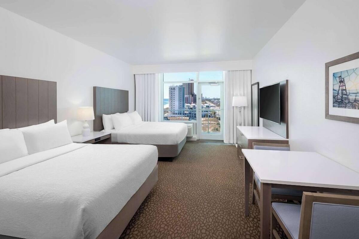 Habitación de hotel con dos camas, televisión, escritorio y vista a la ciudad.