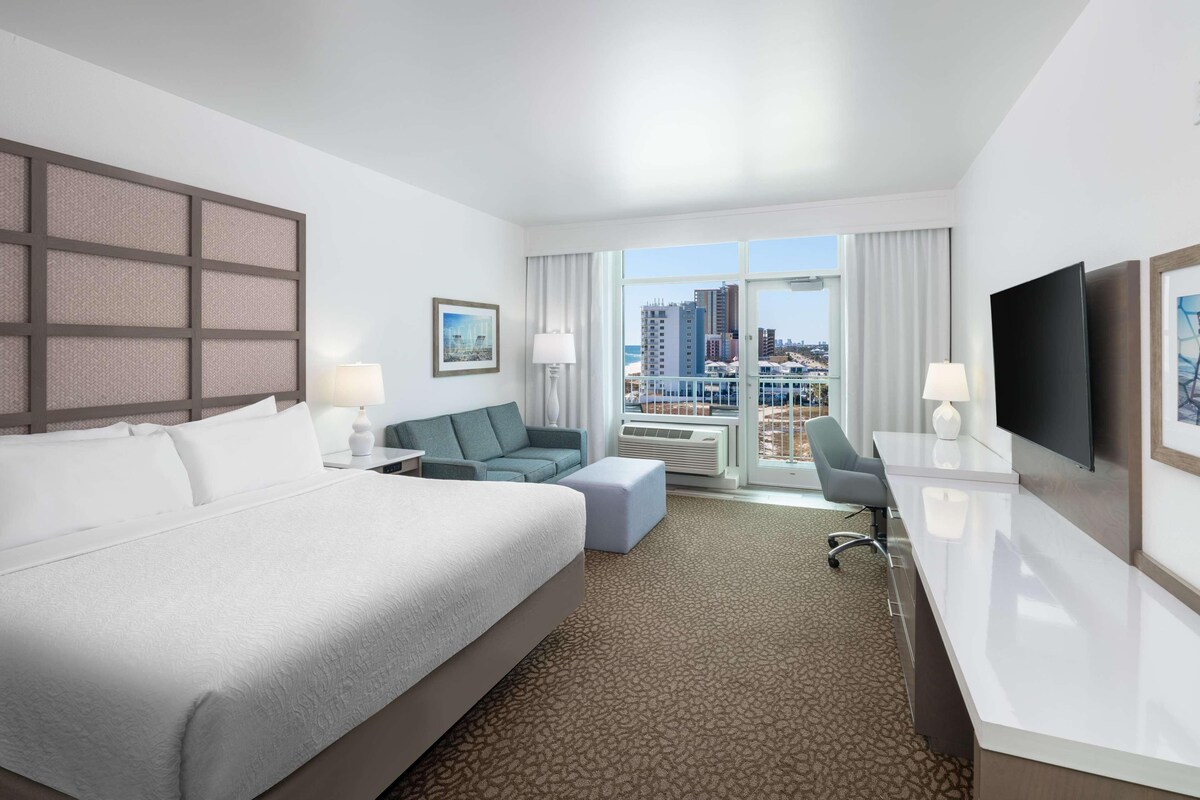 Habitación de hotel con una cama grande, un sofá, un escritorio, un televisor y vistas a la ciudad.