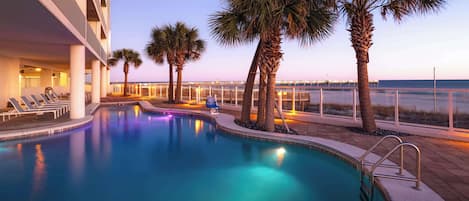 A swimming pool with palm trees, a deck with lounge chairs, and a view of the ocean at dusk.
