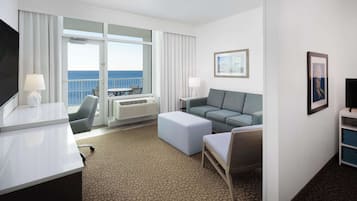 A hotel room with a large window offering a view of the sea, a sofa, a chair, a desk, and a television.