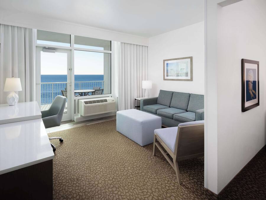Ein Hotelzimmer mit großem Fenster und Meerblick, einer Couch, einem Sessel, einem Schreibtisch und einem Fernseher.