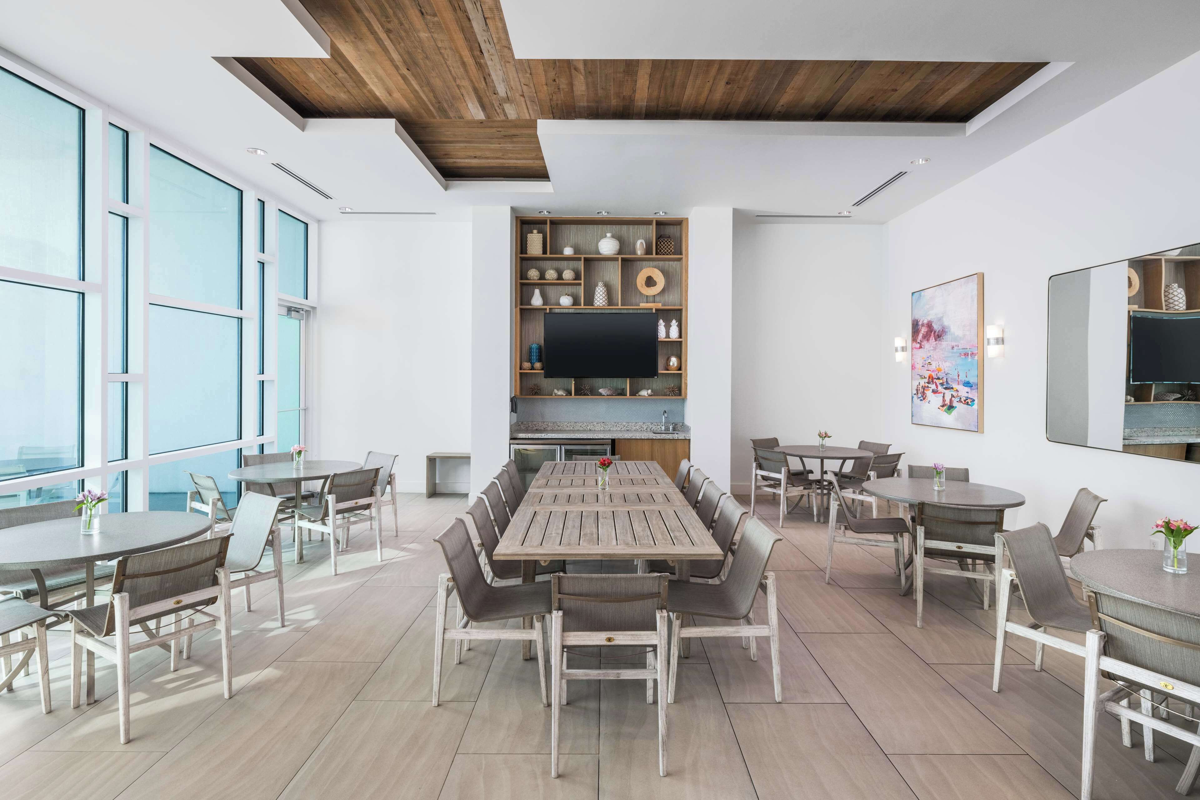 A spacious dining area with a long wooden table surrounded by chairs, a television mounted on a wooden shelf, and large windows providing natural light.