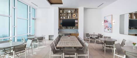 A spacious dining area with a long wooden table surrounded by chairs, a television mounted on a wooden shelf, and large windows providing natural light.