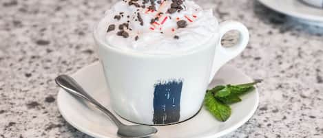 A white cup with a dark blue stripe, filled with a frothy beverage, topped with chocolate shavings and red candy pieces, on a speckled countertop.