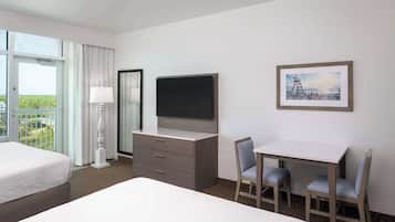 Habitación de hotel con cama, escritorio con dos sillas, televisor, cómoda y ventana con cortinas.