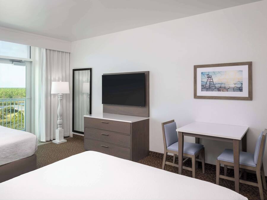 Quarto de hotel com cama, escrivaninha com duas cadeiras, televisão, cômoda e janela com cortinas.