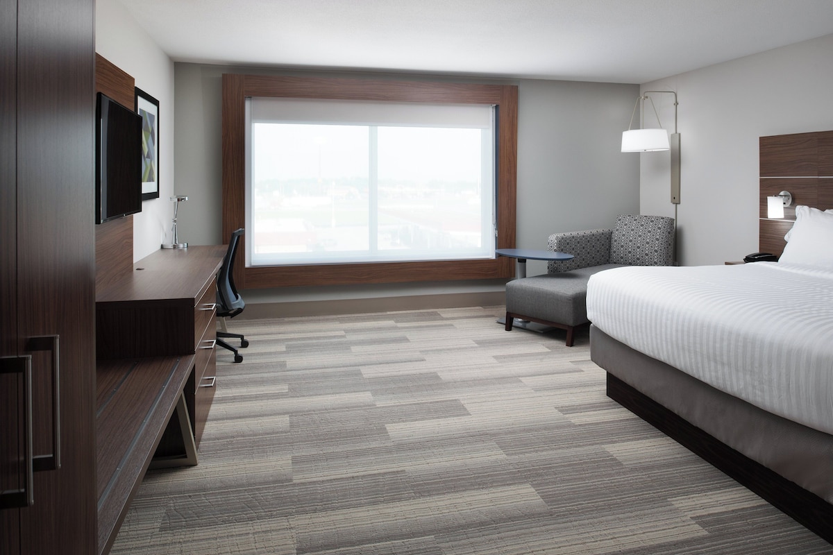 A hotel room with a large window, a bed, a desk with a chair, a small table, and a TV mounted on the wall.