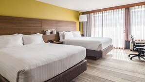 A hotel room with two large beds, a wooden headboard, a nightstand with a lamp, and a view of the outdoors through sheer curtains.