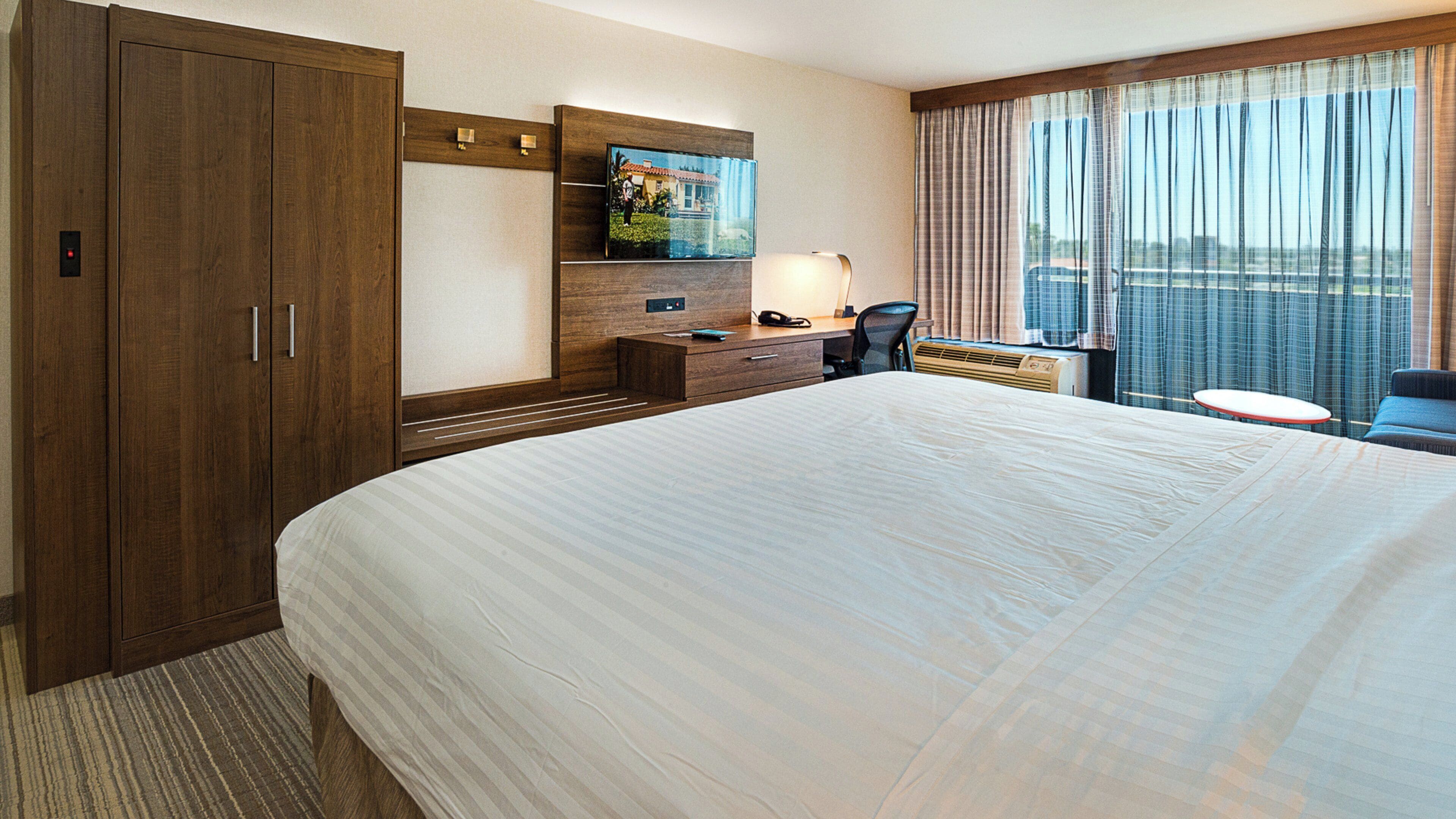 A hotel room with a large bed, a wooden wardrobe, a desk with a chair, a television, and a window with curtains.