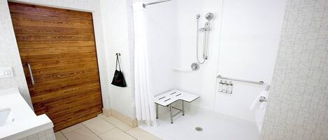 A modern bathroom with a wooden door, a shower with a glass door, a small white bench, and a towel rack.