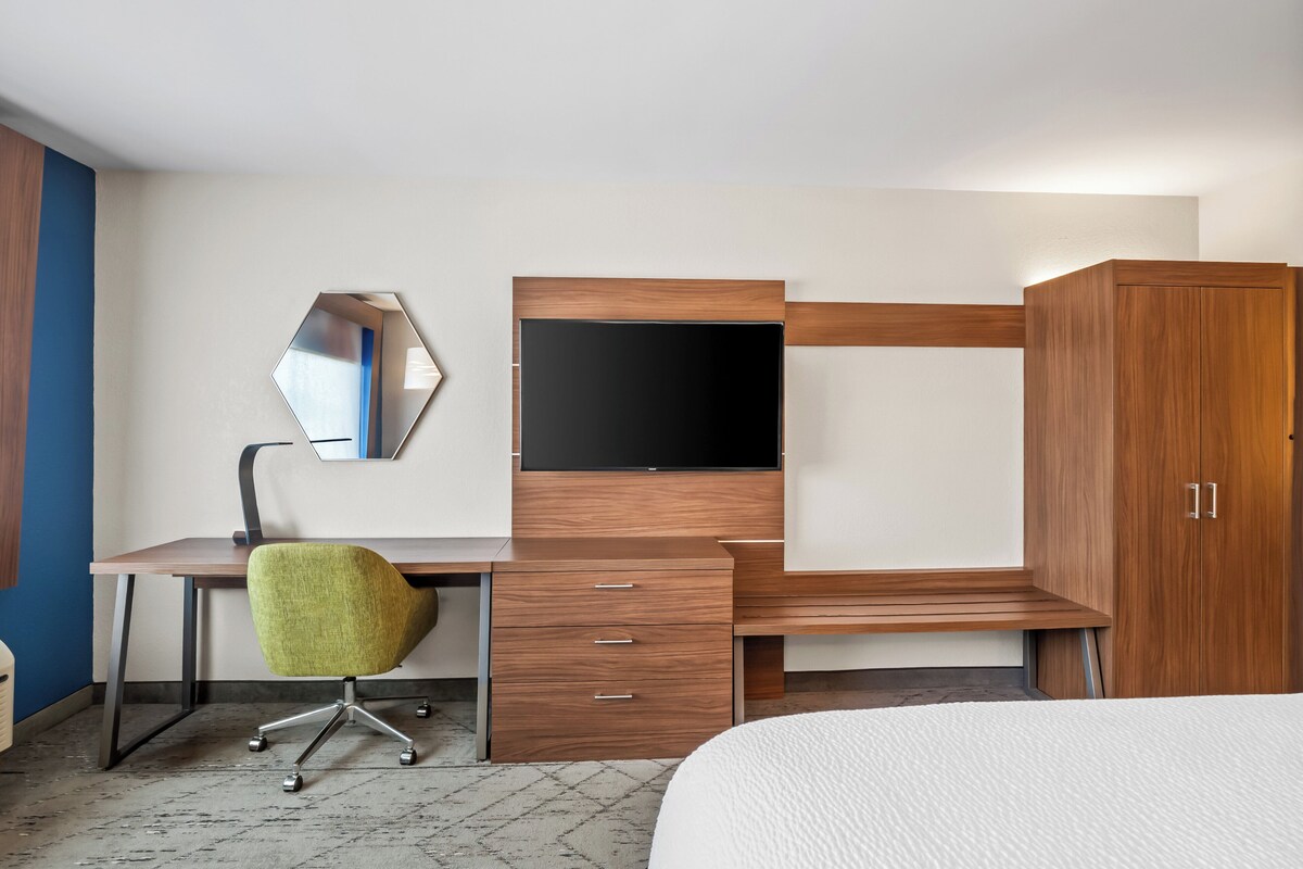 A hotel room with a wooden desk, a green office chair, a flat-screen TV, a wardrobe, and a bed.