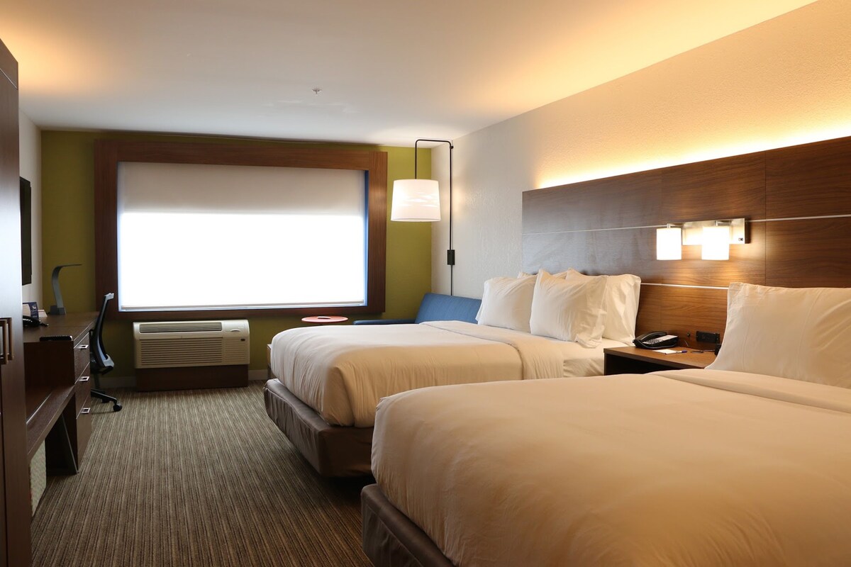 A hotel room with two beds, a desk, a chair, a lamp, and a window.