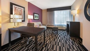 Una habitación de hotel con una mesa de conferencias, un sofá, una ventana con cortinas y una lámpara de pared.