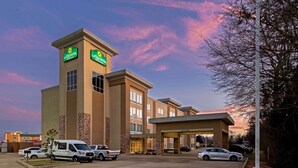 Un hotel con un llamativo letrero de 'La Quinta', un estacionamiento con varios vehículos y un cielo despejado con nubes rosadas.