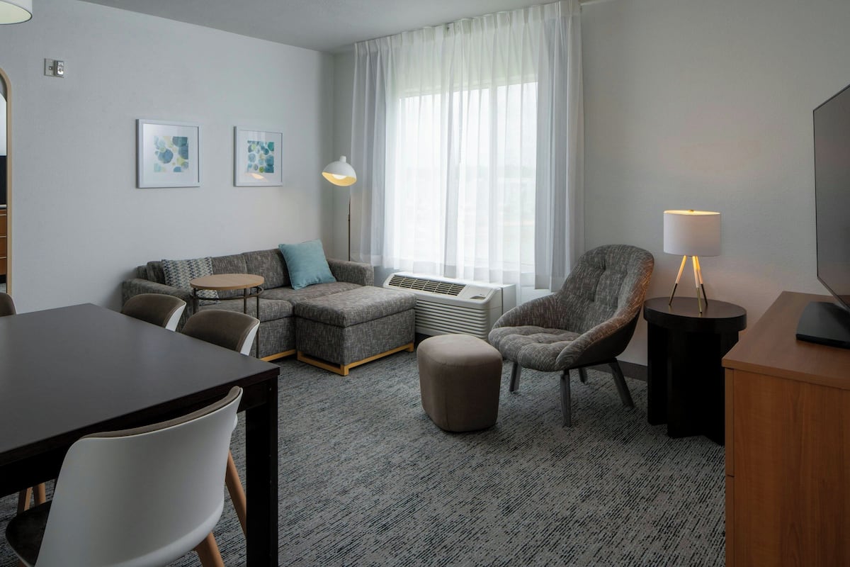 Een moderne woonkamer met een bank, voetenbankje, fauteuil en een eettafel. Er is een televisie, een lamp en kunst aan de muur.