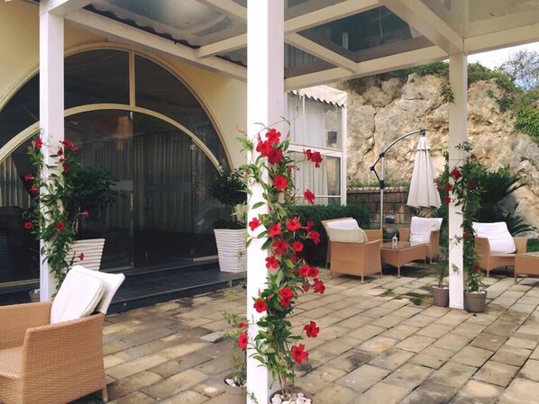 An outdoor seating area with wicker chairs and tables, a stone-tiled floor, and a rock wall backdrop. - San Leucio Resort (Caserta)