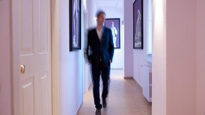 A hallway with framed artwork on the walls, a white door on the left, and a person walking towards the camera. - San Leucio Resort (Caserta)