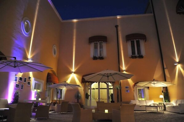 An outdoor seating area with umbrellas, chairs, and tables, surrounded by a building with warm lighting and windows.